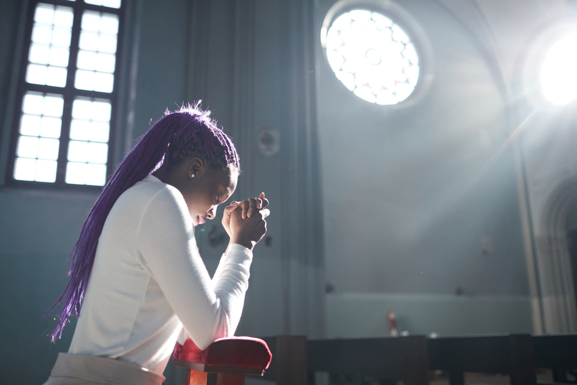 Woman bowing her head with clasped hands in a sunlit church.