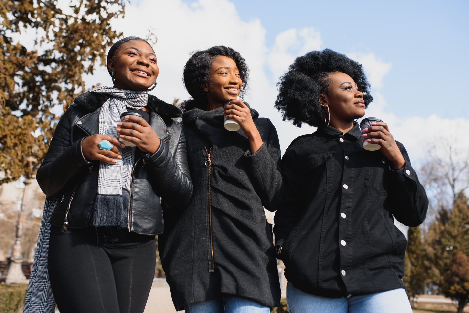 Three women laughing outdoors while holding coffee cups.